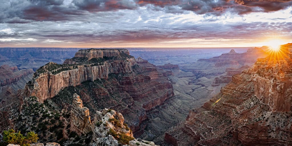 A view over the North Rim of the Grand Canyon. Wotan's Throne, a massive rock formation rises from the floor of the Grand Canyon. It is stair-stepped in nature, with each step having a difference hue. To the right of the image is another canyon wall. Above that is the setting sun giving a flair. The sky is very cloudy with deep reds and purples.